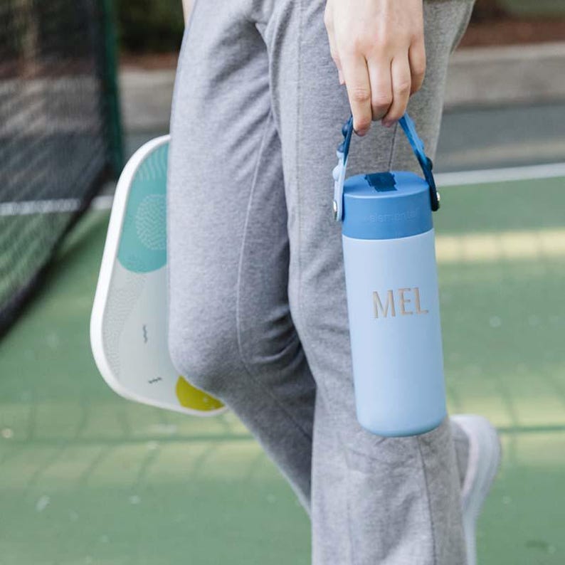 May include: A light blue water bottle with a dark blue lid and handle is held by a person. The bottle has the letters "MEL" printed on it. A white and teal paddle is visible in the background.
