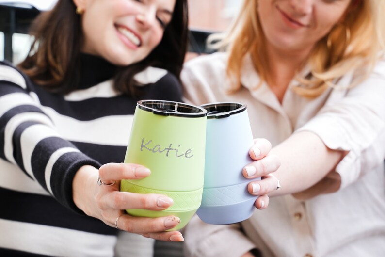 Puede incluir: Dos amigas brindando con vasos de vino aislados de color verde y azul. El vaso verde tiene el nombre "Katie" escrito en &eacute;l.