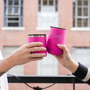 Puede incluir: Dos vasos de acero inoxidable de color rosa con tapas. El vaso de la derecha tiene la palabra "Charity" grabada.