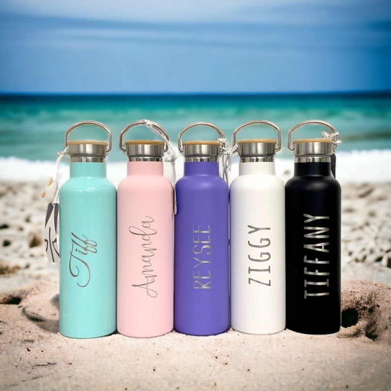 May include: Five stainless steel water bottles with personalized names on a sandy beach. The bottles are in various colors: teal, pink, purple, white, and black.