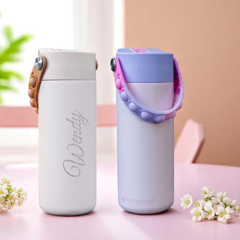 May include: Two stainless steel water bottles. One is white with the name "Wendy" in cursive. The other is light purple with a blue and pink handle. Both have a handle and a lid. The word "elemental" is printed on the bottom of the bottles.