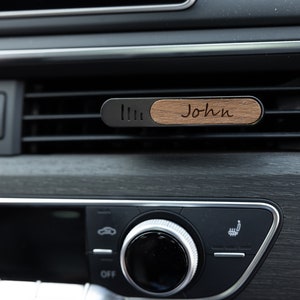 May include: A wooden car air freshener with the name "John" engraved on it. The air freshener is attached to a car's air vent.