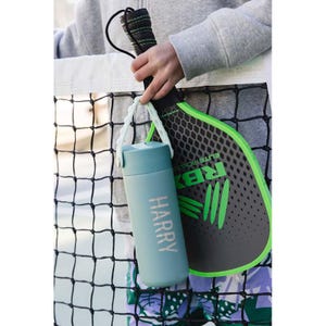 May include: A teal water bottle with the name "HARRY" printed on it, held by a white strap. A black and green pickleball paddle is visible in the background. The paddle has the text "ELITE PICKLE" and the letters "RBX" on it.