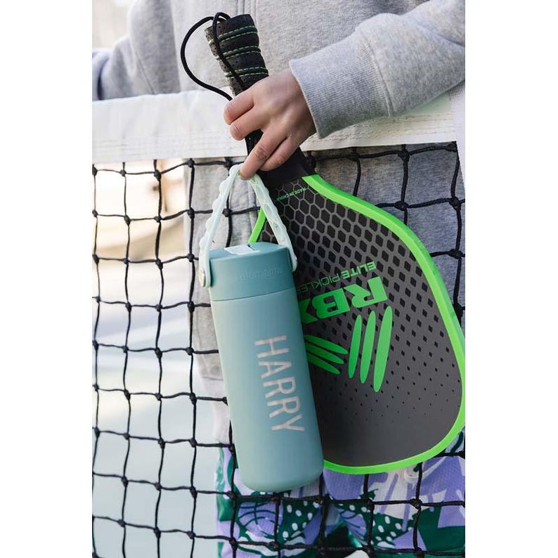 May include: A teal water bottle with the name "HARRY" printed on it, held by a white strap. A black and green pickleball paddle is visible in the background. The paddle has the text "ELITE PICKLE" and the letters "RBX" on it.