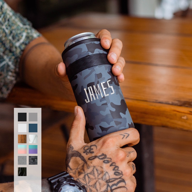 May include: A black and gray camo can cooler with the name "JAMES" printed in white. The cooler is being held by a hand with a tattoo.