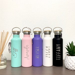 May include: Six personalized stainless steel water bottles with bamboo lids. The bottles are in various colors: teal, pink, purple, white, and black. Each bottle has a name engraved on it:  'Tiff', 'Amanda', 'Reysel', 'Ziggy', and 'Tiffany'.