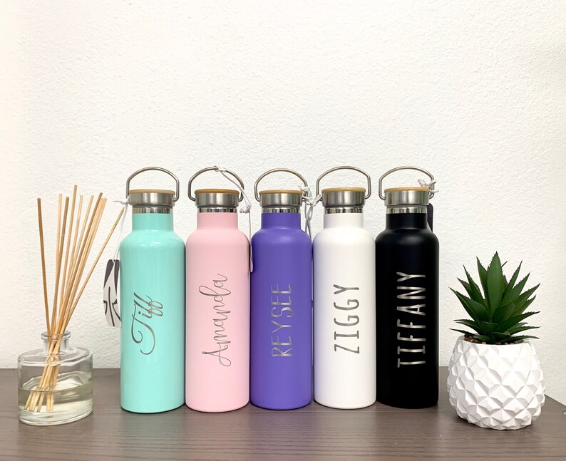 May include: Six personalized stainless steel water bottles with bamboo lids. The bottles are in various colors: teal, pink, purple, white, and black. Each bottle has a name engraved on it:  'Tiff', 'Amanda', 'Reysel', 'Ziggy', and 'Tiffany'.
