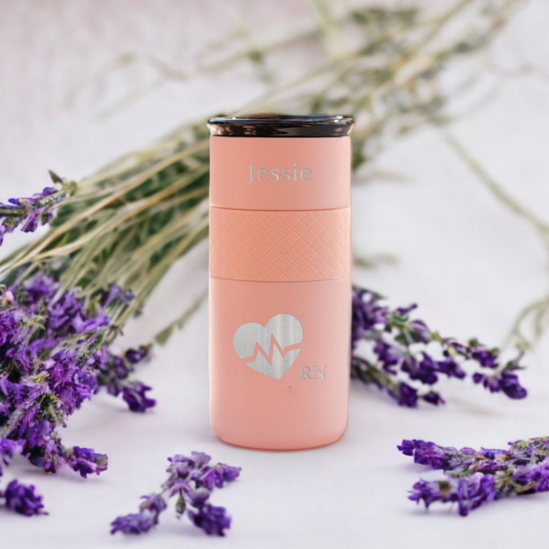 K&ouml;nnte beinhalten: Eine personalisierte Reise-Thermoskanne in Rosa mit schwarzem Deckel. Die Kanne hat ein Herz mit einer Herzschlaglinie und die Buchstaben "RN" eingraviert. Die Kanne tr&auml;gt auch den Namen "Jessie" eingraviert.