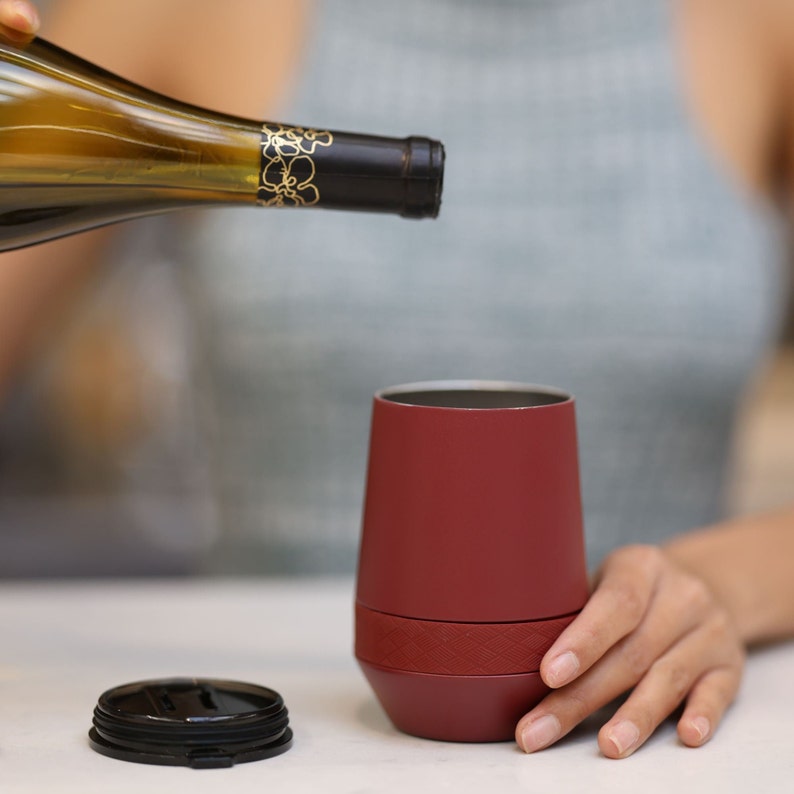 Puede incluir: Una persona vierte vino blanco en un vaso aislante rojo con tapa negra. El vaso tiene una banda texturizada alrededor del medio.