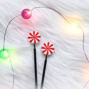 May include: Two black knitting needles with red and white candy cane-shaped tops. The needles are on a white fluffy surface with a string of white lights.