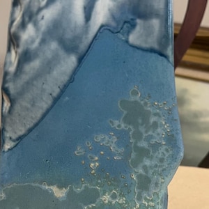 May include: A rectangular ceramic vase with a textured surface. The vase features a blend of blue and white hues, with a unique, organic shape. The glaze has a mottled appearance, adding to its artistic appeal. The vase is a decorative item.