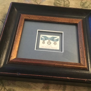 May include: A framed print of a United States Medal of Honor stamp. The stamp features a blue ribbon with three medals and the words "US Navy Medal of Honor".