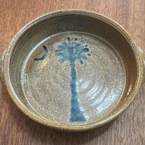 May include: Round, ceramic serving dish with a brown and tan speckled glaze. The interior features a blue palm tree design and a crescent moon. The dish has two handles and is suitable for serving food.