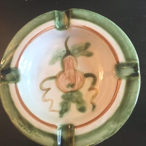 May include: A ceramic ashtray with a green and brown rim and a white center. The center features a hand-painted design of a pear with green leaves.