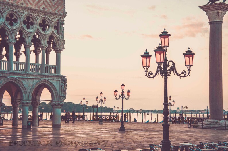 May include: A vintage-toned photo of a grand, arched building with a column in the foreground. The column is topped with a lamp with multiple lights, each with a pink shade. The building has a balcony and ornate details. The scene is bathed in a soft, pink light.