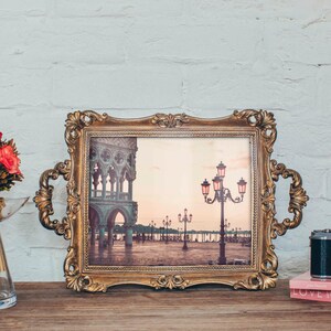 May include: A vintage-style gold-toned picture frame with an ornate design holds a photograph of a cityscape with streetlights. The frame is on a wooden surface with a bouquet of red flowers in a clear glass vase and a camera on a pink book.