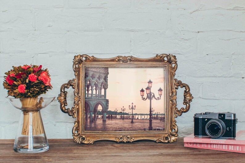 May include: A vintage-style gold-toned picture frame with an ornate design holds a photograph of a cityscape with streetlights. The frame is on a wooden surface with a bouquet of red flowers in a clear glass vase and a camera on a pink book.