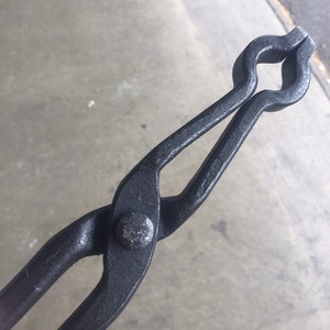 May include: Black metal tongs with a rounded handle and a rivet in the center. The tongs are used for handling hot objects.