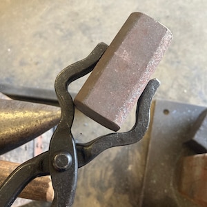 May include: A pair of black metal tongs holding a rectangular piece of metal. The metal is rusty and has a rough texture.