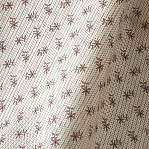 May include: Close-up of a beige fabric with a repeating pattern of small brown flowers and leaves. The fabric is folded over itself, revealing a second layer of the same fabric.