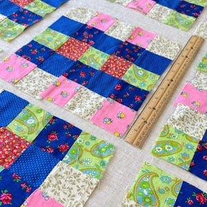 May include: Close-up of colorful patchwork squares in various stages of assembly. The squares feature a mix of floral, paisley, and polka dot patterns in blue, pink, green, and red. A wooden ruler is placed next to the squares.