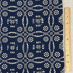 May include: A dark blue fabric featuring a repeating cream-colored geometric pattern of squares and elongated oval shapes. A wooden ruler is positioned vertically along the right edge, showing a length of approximately 12 inches for the visible section.