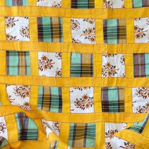 Puede incluir: Un edredón hecho a mano con un diseño de patchwork. Presenta cuadrados alternos de patrones florales y a cuadros en tonos blancos, verdes y marrones, enmarcados por bordes amarillos brillantes. La estética general es vintage y acogedora.