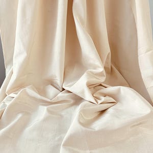 May include: A solid light beige fabric with a subtle sheen and a fine, woven texture. The material is draped in soft, vertical folds, creating deep shadows and highlights across the surface, emphasizing the smooth, consistent color throughout.