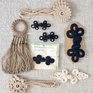 May include: Assortment of handmade beige and black frog fasteners, including a packet of black frog fasteners labelled "HAND MADE FROG FASTENER STYLE NO. AF 973 COLOR", and a beige tassel with beads.
