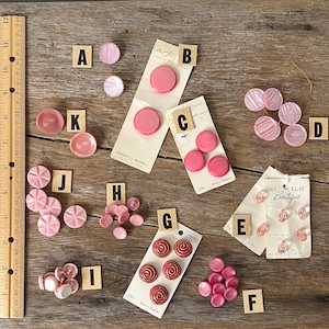 May include: Assortment of pink buttons in various shapes and sizes, including round, swirl, and striped designs. Some buttons have a white center. The buttons are arranged on a wooden surface with price tags and labels.