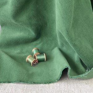 Puede incluir: Primer plano de una tela verde texturizada con tres carretes de hilo verde. La tela está drapeada, mostrando pliegues y un borde sin rematar. Los carretes de madera tienen etiquetas y son de varios tonos de verde.