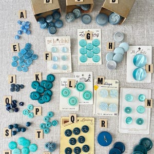 Puede incluir: Surtido de botones vintage en varios tonos de azul y turquesa. Los botones están dispuestos en orden alfabético sobre un fondo de tela gris claro. Algunos botones están en tarjetas con etiquetas, incluyendo "Le Chic", "La Ging" y "Lansing".