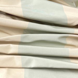 May include: Close-up of folded fabric with a striped pattern. The fabric features alternating bands of light beige, pale green, and off-white. The material appears to be smooth and possibly silk or satin, with subtle folds and highlights.