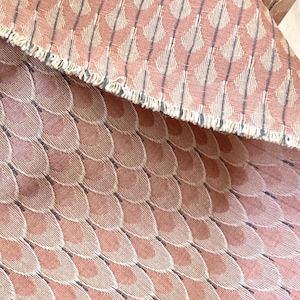 Może przedstawiać: Zbliżenie tkaniny z powtarzającym się wzorem nakładających się, półkolistych kształtów w odcieniach różu i beżu. Wzór jest zaakcentowany pionowymi liniami w ciemniejszym odcieniu. Tkanina ma postrzępioną krawędź.