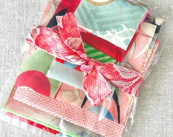 May include: A collection of colorful fabric scraps in various patterns, including floral and geometric designs, tied with a red patterned ribbon. The fabrics are packaged in a clear plastic bag, ready for crafting or quilting projects.