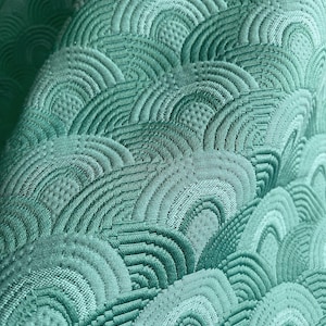 Puede incluir: Una tela de color verde menta con un patrón en relieve de semicírculos superpuestos. La textura consiste en líneas cosidas en relieve que forman arcos con un fondo punteado dentro de cada arco, creando una superficie con dimensión.