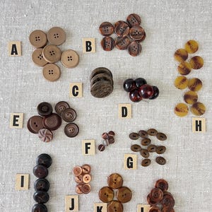 May include: Assortment of brown, black, and tan buttons in various shapes and sizes. The buttons are labeled with letters A through L.