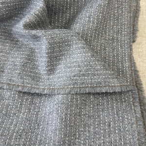 May include: A grey fabric featuring a pattern of vertical white dashed lines. The material has a soft, textured surface with a visible nap and a finished edge that shows a thin brown border detail along the hemline of the folded textile piece.