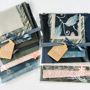 May include: Two packages of fabric scraps in shades of green, blue, and pink. The fabric scraps are wrapped in clear plastic and tied with a gray ribbon. Each package has a brown tag with a white bird design.