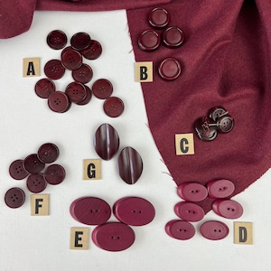 May include: Assortment of burgundy buttons in various shapes and sizes, including round, oval, and rectangular. The buttons are arranged on a white surface with letters A through G labelling each group.