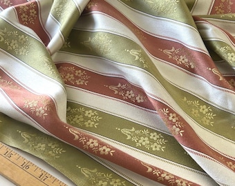Peut inclure: Gros plan sur un tissu plié avec un motif à rayures. Les rayures alternent entre le blanc, le rose poudré et le vert olive. Des motifs floraux dorés ornent les rayures roses et vertes. Une règle en bois avec des mesures en pouces est au premier plan.