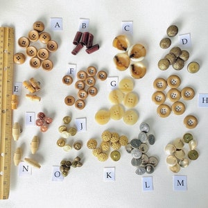 May include: Assortment of buttons in various colors and materials, including brown, beige, gold, and silver. The buttons are arranged in groups labeled with letters A through M.