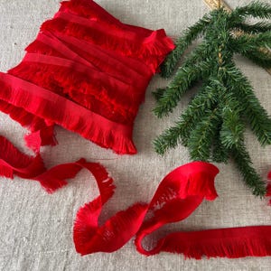 Puede incluir: Cinta roja brillante con flecos, dispuesta artísticamente, con una apariencia lujosa y texturizada. La cinta se muestra junto con una ramita de pino verde, lo que sugiere un tema festivo o artesanal. Los bordes de la cinta están acabados con una suave franja.