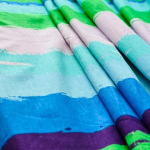 May include: Striped fabric with a colorful abstract design. The fabric features alternating stripes of green, purple, blue, and light blue. The stripes are painted with a brushstroke effect.