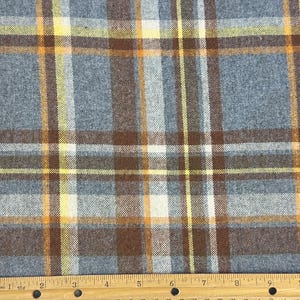 May include: A woven fabric featuring a plaid pattern in shades of slate blue, dark brown, white, and bright yellow. A wooden ruler is positioned at the bottom, showing the fabric width spans approximately 9.5 inches across the visible section.