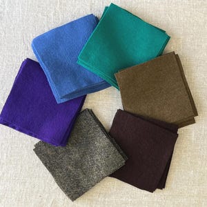 May include: A collection of square felt fabric pieces in various colors, including blue, teal, brown, purple, and a tweed-like gray. The felt squares are arranged in a circular pattern on a neutral-colored surface.