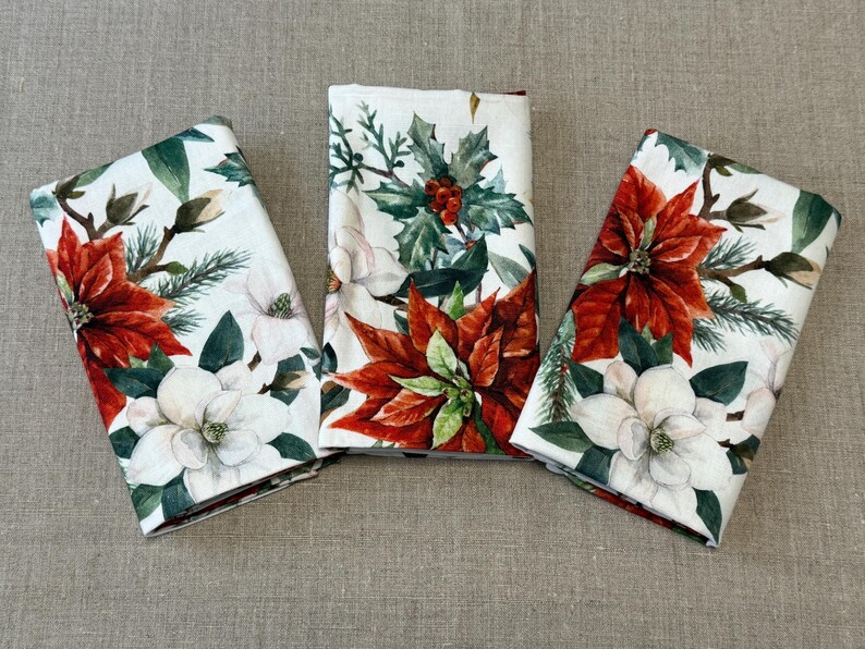 May include: Three white linen napkins with a floral design featuring red poinsettias, white magnolia blossoms, and green holly leaves.