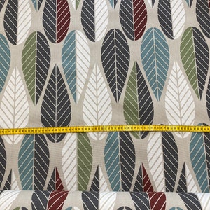 Op de afbeelding: Beige stof met een herhalend patroon van gestileerde bladeren in tinten blauw, groen, zwart en rood. De stof wordt gemeten met een gele meetlint.