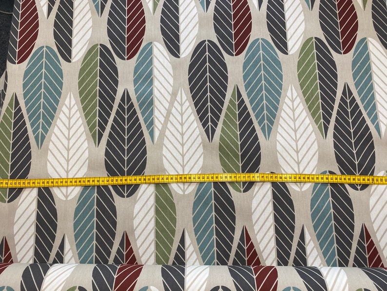 Op de afbeelding: Beige stof met een herhalend patroon van gestileerde bladeren in tinten blauw, groen, zwart en rood. De stof wordt gemeten met een gele meetlint.