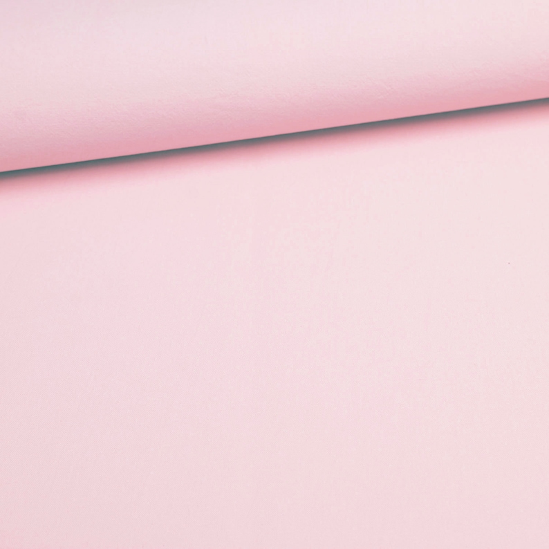 May include: A close-up of a roll of light pink fabric. The fabric is smooth and appears to be a solid color. The top edge of the fabric is rolled over, creating a slight shadow.